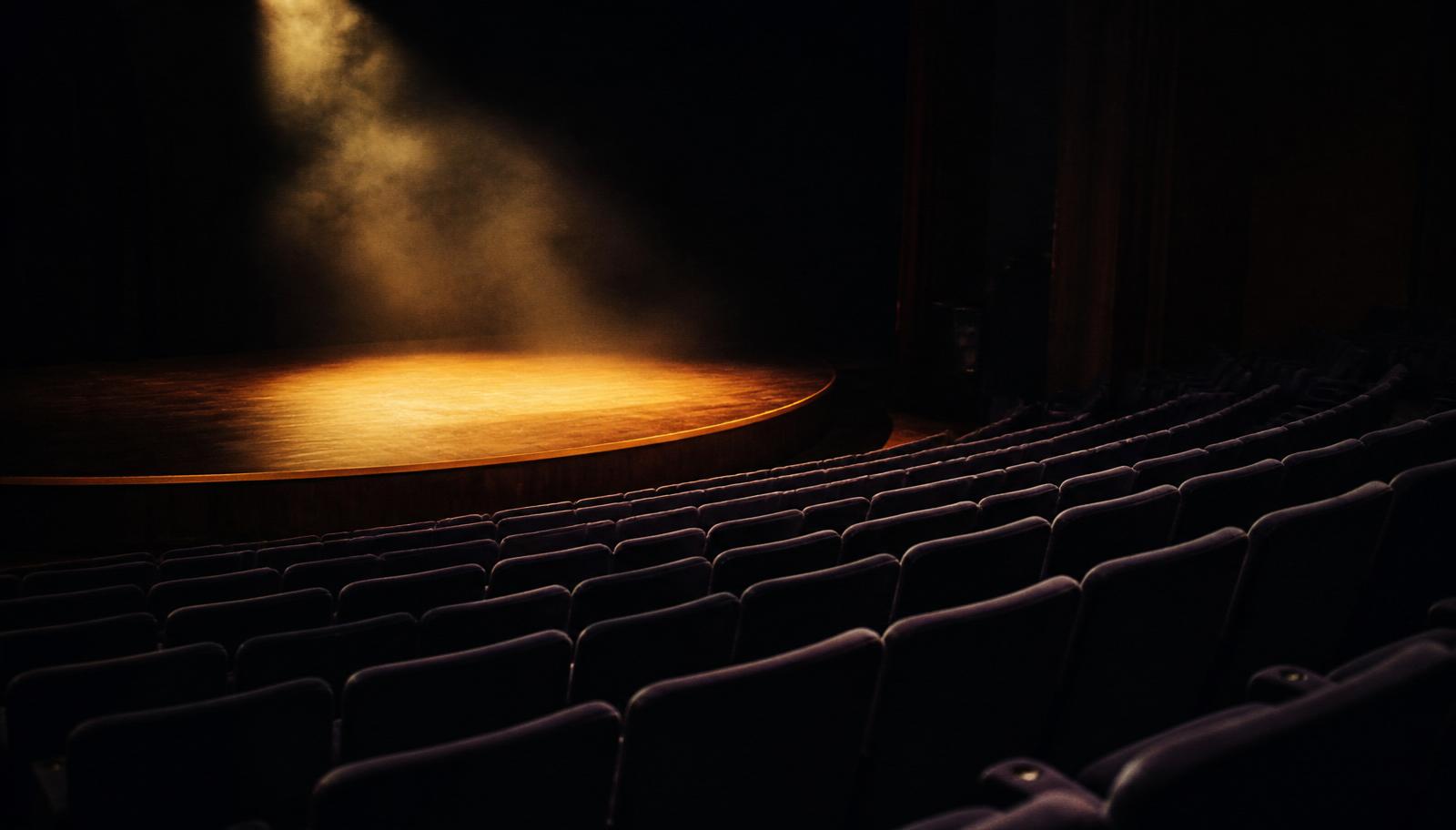 An empty auditorium in dim light, a single warm spotlight resting on a wooden stage before a talk.